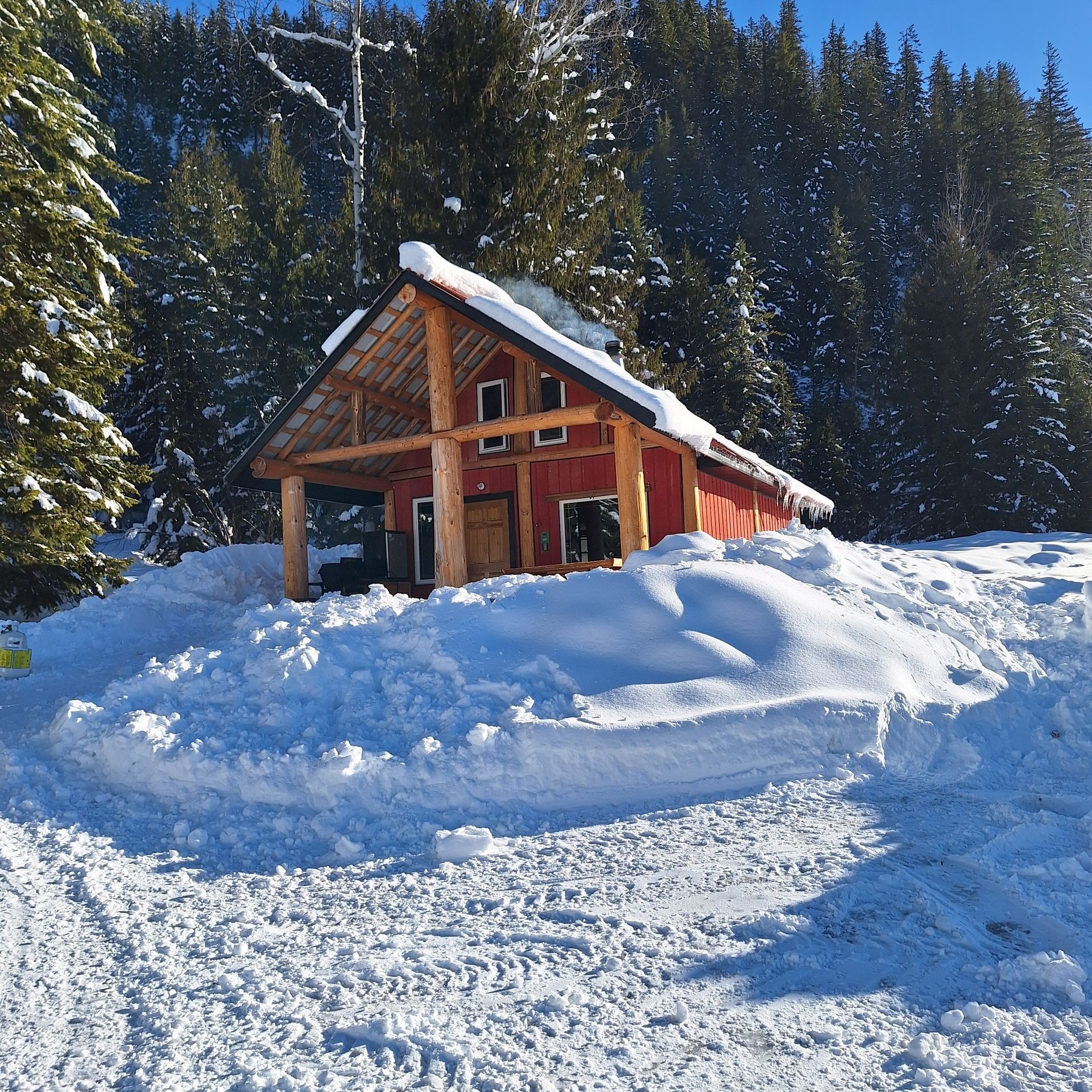 Red wooden cabin in a snowy forest with snow-covered roof and trees under clear blue sky.